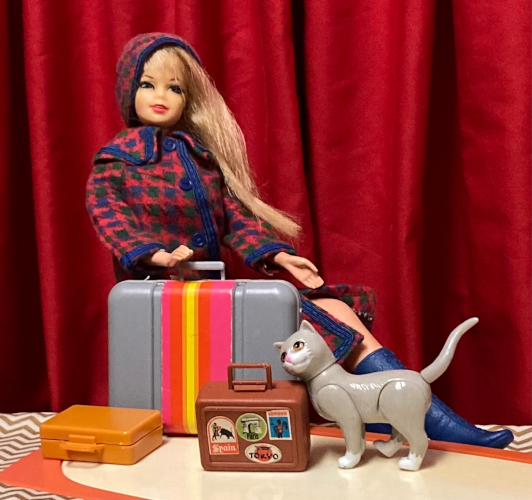 Vintage blonde Stacey wears Mainly for Rain, a red, green and blue houndstooth long coat with blue trim and buttons, matching hat, and high blue boots. She is seated--on what, we can't see--behind a 1970s plastic coffee table of pink with a white oval top. Before her are three "suitcases." The silver-look plastic suitcase stands upright, partly balanced by her hand. It has a pink, yellow and orange stripe down the middle somewhat reminiscent of a luggage strap, but wider. This looks like a fine suitcase or at least briefcase size for Stacey, from this angle. On its side lies the gold- or mustard-colored case from the Friend Ship, which, in Stacey scale, is about the size of a dictionary. Standing next to it is a brown case, about the size of a lunch box, with stickers for four locations including Tokyo and Spain. Unlike the foregoing hat boxes, these are actual stickers with color illustrations. Kiki stands on the table and rubs her cheek against the corner of the brown case.