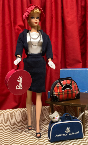 Commuter Set reproduction Barbie with blonde ponytail, bead necklace, red feathery crescent hat, white satin blouse, white gloves, and navy jersey Chanel-style suit stands holding a red cardboard hat box with the Barbie signature in white on the front. Next to her is a small wood table (Mattel Modern) holding the plaid overnight bag with black trim from Winter Holiday. On the floor before them sits the American Airlines flight bad, unzipped. Kiki's head and tail emerge from the AA bag.