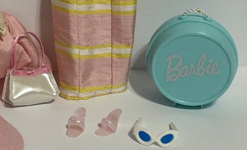 Photo from eBay listing shows the bottom part of a doll's sheath dress, pink with yellow and white stripes in a high-sheen fabric. Next to the dress is a pale greenish blue hard plastic hat box with the Barbie logo in white and a white cord handle. Other pieces are white cat eye sunglasses with blue lenses looking a little melty--less sharp than the vintage ones--along with pearlescent pinkish open-toed shoes and a pearlescent white purse with pink trim. A high-sheen pink jacket sits just on the edge of the frame and may have a flower corsage on the lapel. If not wedding planner I guess this style could be Mother of the Bride.