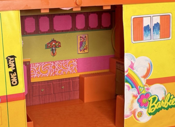 Color photograph. The sliding door of the camper stands open, revelaing an interior wall lined with pink-and-woo-grain cabinets (screened on), a tiffany wall lamp, a poster of a neon-colored owl, and a pink-and-orange paisley backsplash or wallmounted cushion (it's all just printed on). 