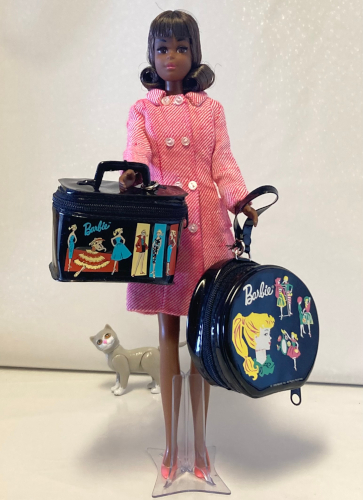 On white vinyl backdrop, Black Francie reproduction stands wearing Shoppin' Spree. In one hand she holds a rectangular zippered black vinyl train case covered with screened-on images of vintage Barbie modeling her fashions before colorful rectangles. In the other she holds a sizeable hat box with a closeup of Barbie's profile and a handful of small images of Barbie in assorted fashions, also on black vinyl and closed with a zipper. At the back of the scene Kiki, wandering by, looks over curiously.