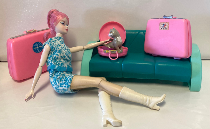 On a stark white vinyl backdrop, Proudly Pink Silkstone Barbie, wearing a blue-and-white Seventies Glamour Group minidress with wide blue belt and Seventies calf-length pak boots, sprawls on the floor next to a hard plastic green sofa that is 90s Barbie but actually kinda child-sized relative to Barbie. Behind her is a large Samsonite-style suitcase in bright pink with metallic painted accents along the top edge where the case closes and locks and where the handle attaches. On the front of the suitcase, centered near the top, is a sticker with a blue Pan Am logo. On the small couch sits a slightly smaller suitcase of the very same design, but with a United logo sticker in red and blue. Next to that a pink plastic hat box lies open, with the same metal  appearancce and the site of the hinged handle and molded locks. The lid of the hat box is open at a 90 degree angle and Kiki sits inside, fitting nicely. It looks like a good size for her to curl up comfortable inside, though now she sits erect. She looks back over her shoulder at Barbie, who reaches out to pet her.