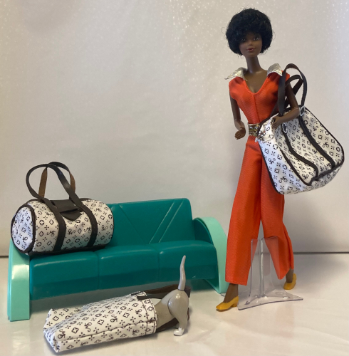 On a stark white vinyl backdrop, 1980 Black Barbie in a late-Seventies Best Buy orange jumpsuit with gold accents stands next to the diminutive hard plastic couch. Over her arm is a sizeable cloth suitcase in white festooned with little logos and monograms and with leather straps and piping. On the couch, nearly dwarfing the couch, is a cylindrical duffel. On the floor before the couch a rectangular tote lies on its side. Kiki's hind legs and tail are visible sticking out from the tote as if she is playing.
