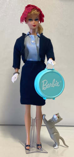 On a stark white cinyl backdrop, blonde ponytail Barbie reproduction wears her own repro Commuter Set suit, now with the blue-and-white checked collared shirt and no necklace. In one hand she holds a hard plastic teal/aqua hat box by its white cord. Kiki stands beside her, stretched with front paws up on her leg, gazing toward the hat box curiously or longingly.