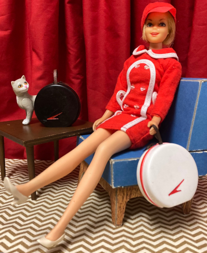 Blonde Casey wears a red minidress/jacket with white trim and a double row of white buttons with a matching red hat plus a pair of white closed-toed heels. She sits in the blue chair, holding Tammy's hat box. A red V-shaped swoosh or boomerang decorates the front of the hat box. The wooden table, behind her, holds the black version of the hat box sitting upright, showing the same red swoosh. Kiki sits behind the second hatbox and looks back over her shoulder toward Casey.