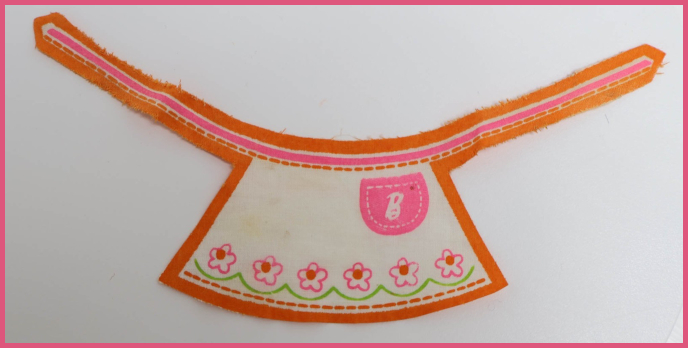 Photograph of doll apron cut form a single piece of fabric printed with an orange outline, pink flowers with green leaf-like underlining, and a pocket-shaped pink bit emblazoned with Barbie's script B.