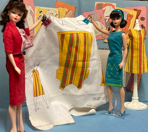 Back in the fashion design studio diorama, the fashion designer "dolls" hold up a sheet of white woven fabrin on which are printed the pieces of a shift dress with vertical stripes in yellow, green and red, and lables like "DRESS B FRONT"; "HEM". A character about 3" high or so is also printed on the fabric, shown wearing the dress. The illustration's dress is in color like the garment pieces but she's otherwise a black line drawing. In the background of the figure, the white dressform stands with a constructed example of the shift dress displayed on it.
