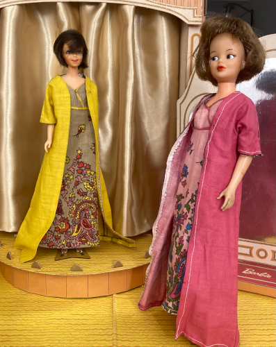 Photo taken in the interior of the vintage Barbie's Fashion Shop, as many of the remaining images in this post will be. Brunette Casey stands on the stage wearing a floorlength yellow coat that spreads around her feet, along with a long taupe dress with paisley print (this item was also shown in a Mercari listing earlier). Note, the stage's curtains are normally white vinyl, like a shower curtain. I needed replacements and made them from an antique gold satiny lining fabric which I find a little too reflective in these images, so they're not as nice as they could be. Anyway, in the foreground is the later-model Tammy, smaller than original Tammy and with bobbed brown hair and rubber arms. She wears the same outfit, only her jacket is rose pink and her dress a pale pink with the paisley pattern in blue, green, and rose pink. The Fashion Shop's three-way mirror is behind her and reflects her back, blurrily.
