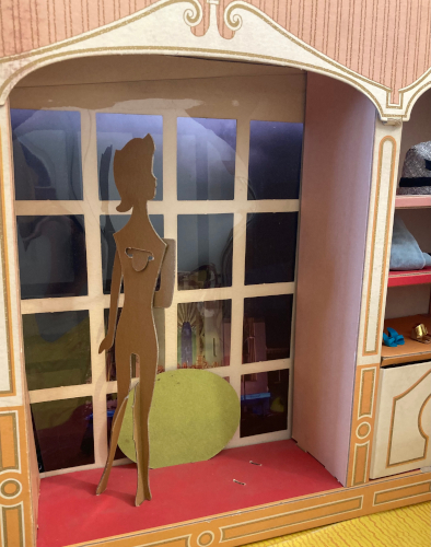 A cardboard silhouette of a ponytailed female figure standes before a darkened window of cellophane crisscrossed by off-white chipboard to form panes.