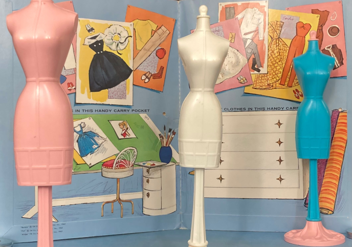 Three Barbie-sized plastic dress forms stand in a row, one pink, one white, and one blue, in front of a paper doll folder that stands propped up, illustrated with scenery from a fashion designer's studio.