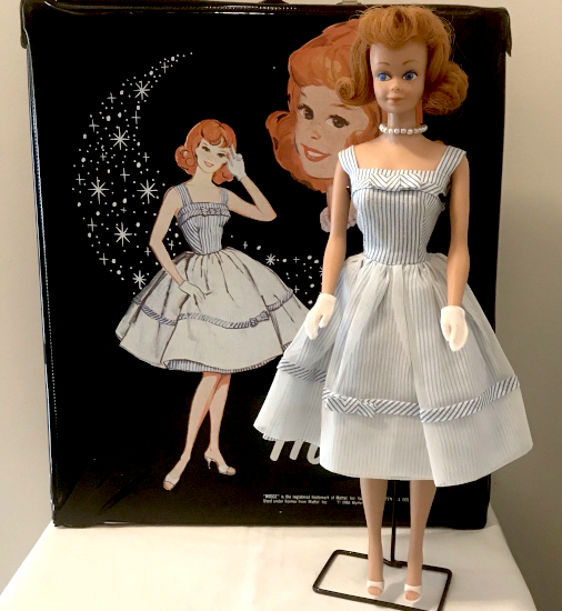 Color photograph. Vintage Midge wirth red flip hair on a doll stand, wears Movie Date fit-and-flare, blue-and-white striped dress with sheet overskirt, pearl necklace, short white gloves, and white open-toed shoes. In the backgorund is a black vinyl doll carrying case with an illustration of Midge wearing exactly the same thing sans necklace. A larger Midge head floats at her shoulder and a trail of sparkles dashes across the background.