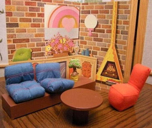 A warm, handsome room consisting of a back wall of vari-colored brick with a graphical heart painting, an orb wall sconce, Malm-type fireplace, screened-on credenza topped by flowers, vases, and green throw pillow. In the foreground is a brickish brown couch topped by denim pillows, a stuffed orange chair with no arms, and a round circular table in the same plastic as the couch. One end of the couch is free of cushions to make a size table on which a little potted tree rests. This scene is set atop a wood-textured surface which isn't part of the Mattel set but adds to the warmth.