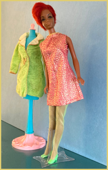 Color photograph. 1960s Julia doll with very oxidized and rather puffy pixie haircut wears the salmon-and-gold minidress from Velvet Venture, which has an A-line shape and a thin strip of goldtrim at the neck. Shewears acid-green hose and bright green, square-toed shoes. On a teal-colored mannequin beside her is the Velvet Venture jacket, also acid green, in a velvety material, the same length as the dress, long-sleeved with gold braid trim at the wrists and outlining the cream-colored lapels. At the waist are two gold seed beads connected by short gold chain. The background is light blue with a dark teal floor beneath them.